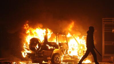 A photograph of a street riot, with a person walking by a burning car.